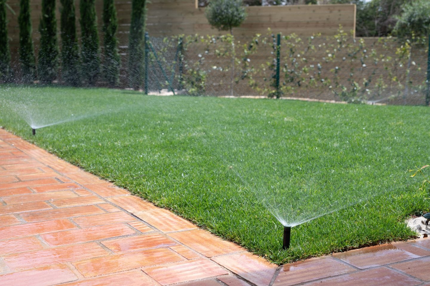Lawn sprinklers watering a lush garden with synthetic grass next to a wet, red-tiled pathway. A fence and trees are visible in the background.