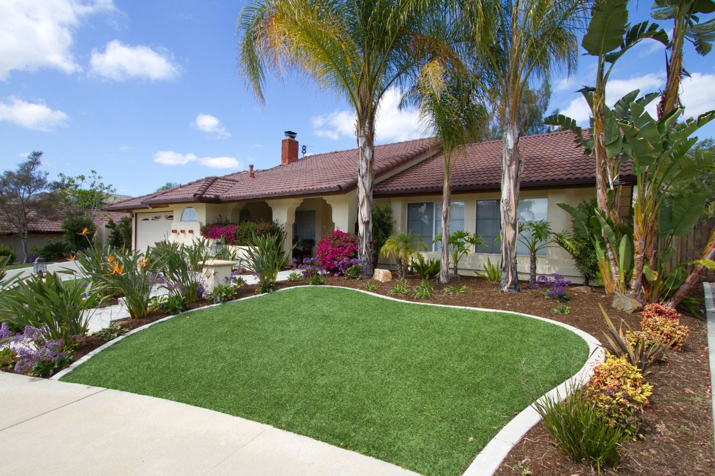 A charming single-story house with a red-tile roof is nestled amid palm trees and a beautifully landscaped lawn of vibrant flowers and synthetic grass. All set against a bright blue sky with scattered clouds in the background.