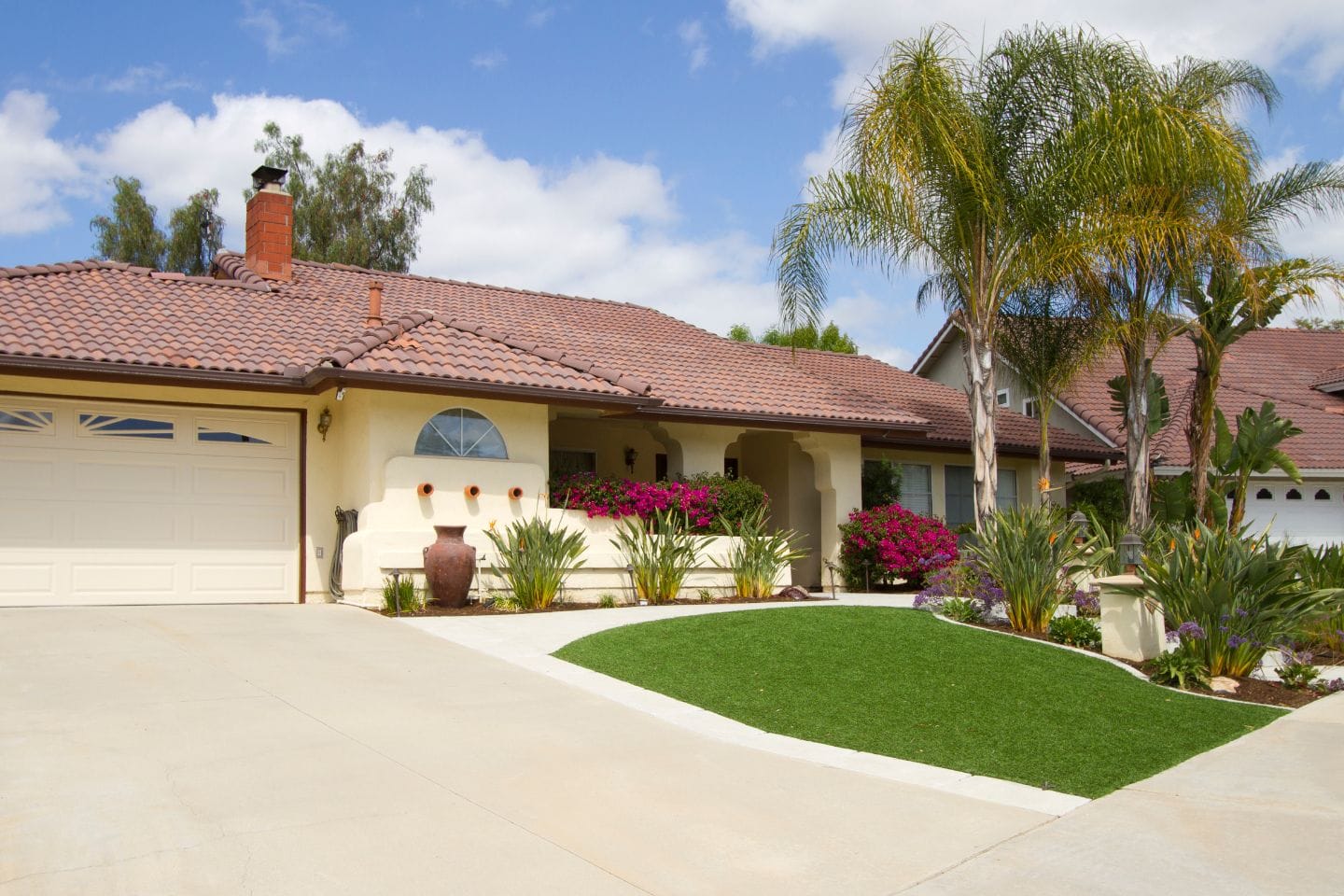 Single-story house with tan walls, red-tiled roof, and a garage. The landscaped front yard, featuring palm trees and vibrant flowers, is complemented by synthetic grass for easy maintenance. Potted plants add charm to this delightful home.