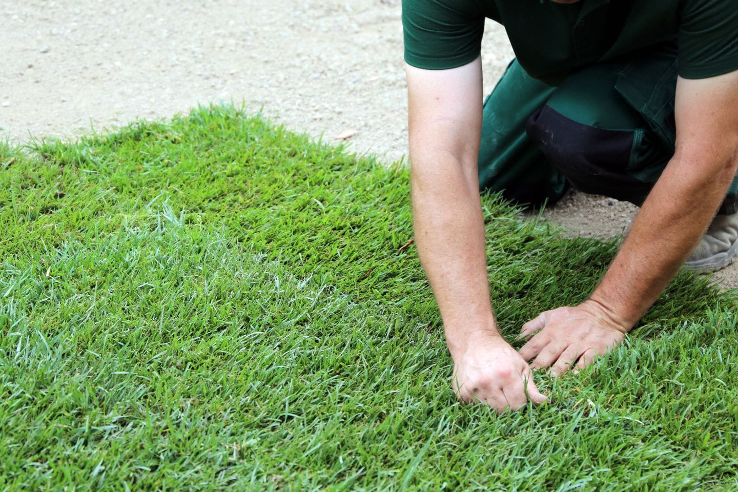 A person kneeling and laying synthetic grass on a dirt surface, carefully positioning the patches to fit seamlessly.