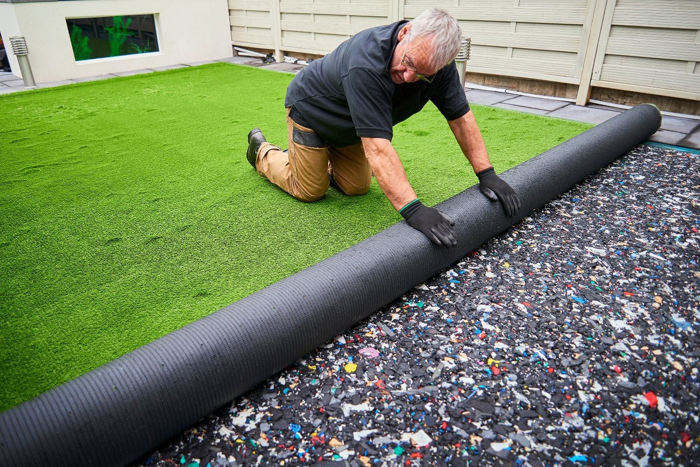 A man in gloves kneels on synthetic grass, unrolling a large sheet over a colorful textured base.