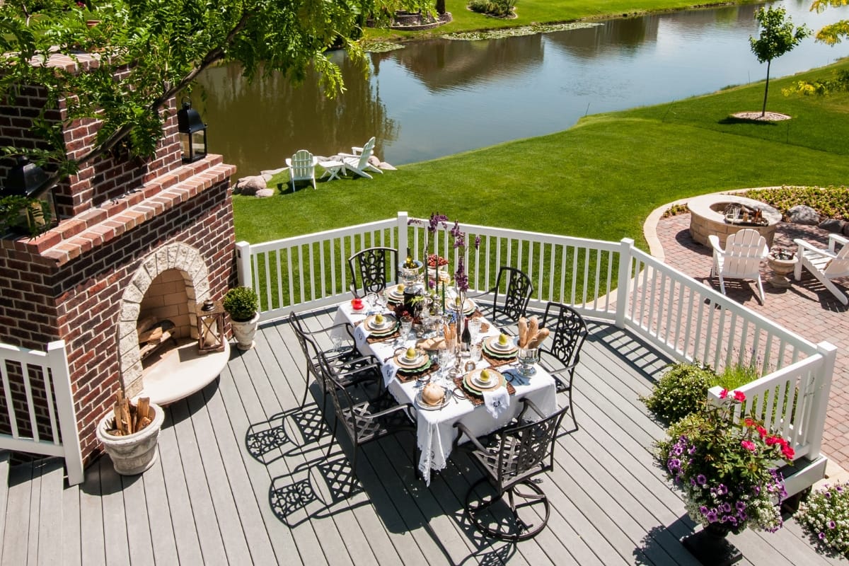 A wooden deck with a brick fireplace and chimneys overlooks a lake, featuring an outdoor dining table set for a meal. The hardscaping services in Atlanta deck also includes potted plants and offers views of a grassy yard with a fire pit and lawn chairs.