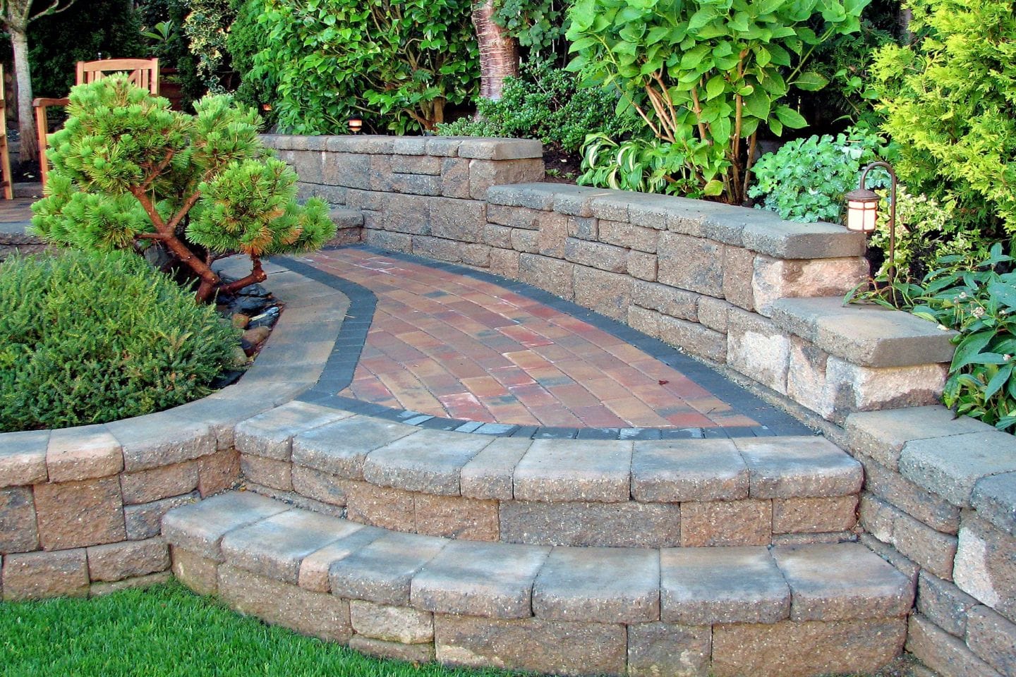 Garden pathway with red and brown bricks, bordered by stone walls, surrounded by green plants and shrubs.