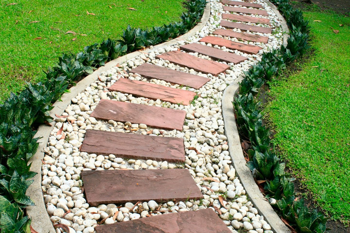 A curved garden path with brown stone slabs is set in white pebbles, bordered by green grass and small plants.
