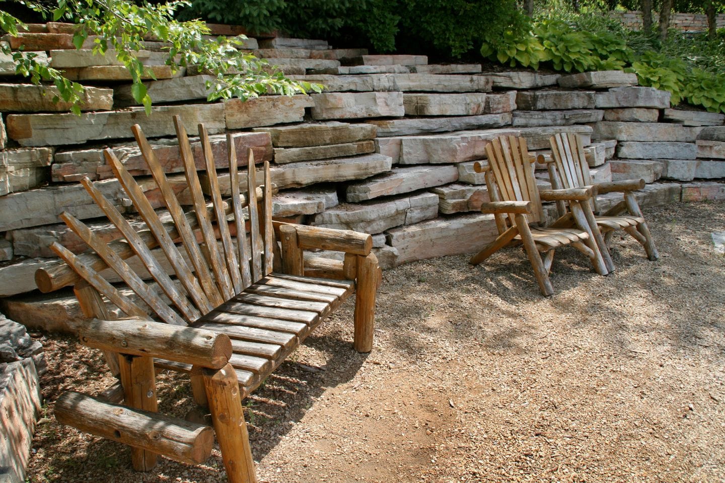 Two wooden chairs, one with a back made of vertical logs, are placed on a gravel surface in front of a stone wall in a garden setting.