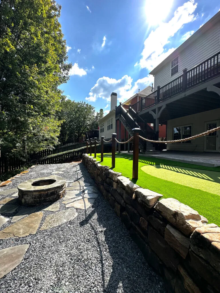 Backyard with a stone walkway, fire pit, artificial grass, and a raised deck. Sunlight casts shadows from nearby trees and a building.