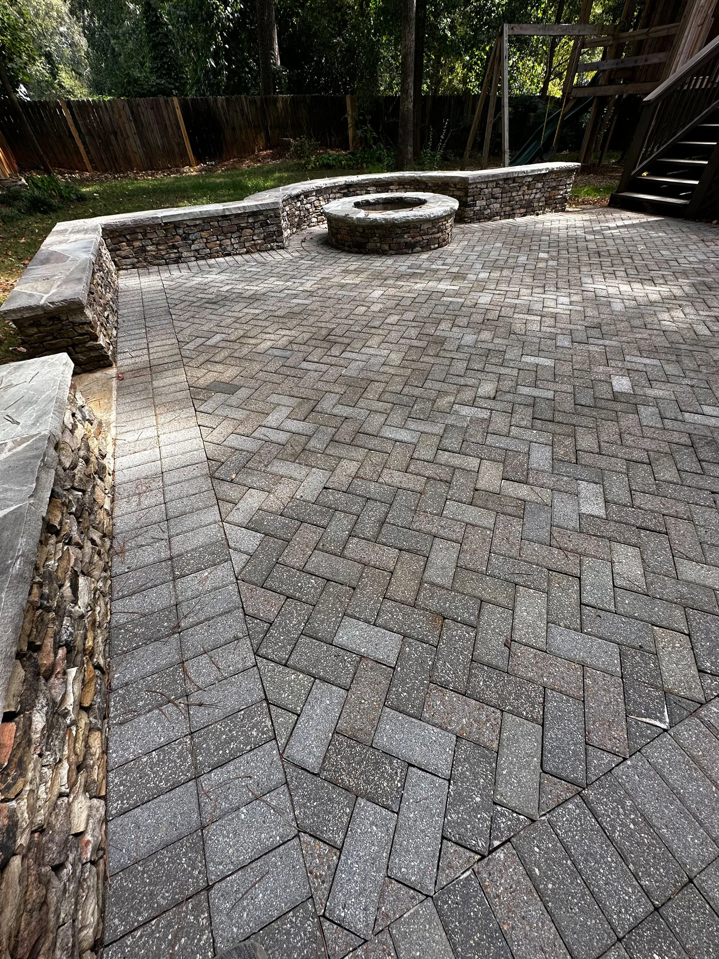 A paved patio with a herringbone pattern, featuring a circular stone fire pit and surrounding stone seating wall, set in a backyard with a wooden fence and trees.