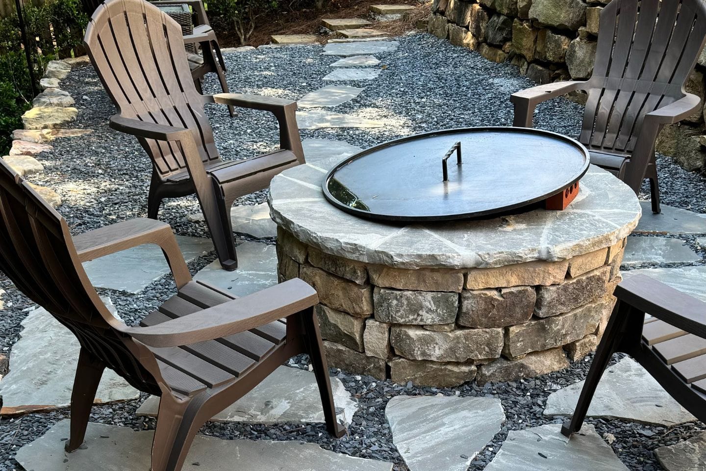 A patio crafted by Atlanta hardscape contractors features four wooden chairs encircling a round stone fire pit with a metal cover, all set on a flagstone and gravel floor.
