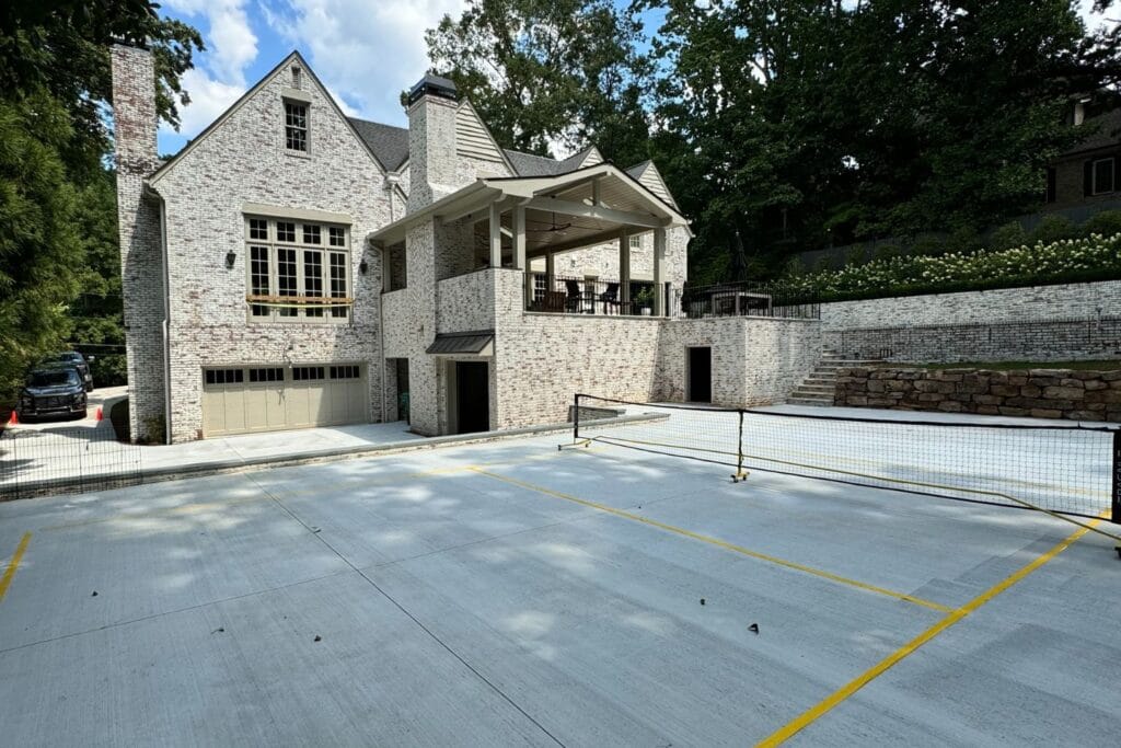 Large brick house with outdoor patio crafted by top Atlanta hardscape contractors and a convenient garage entrance. The property features a concrete area lined for pickleball or tennis, surrounded by lush trees and a charming stone wall.
