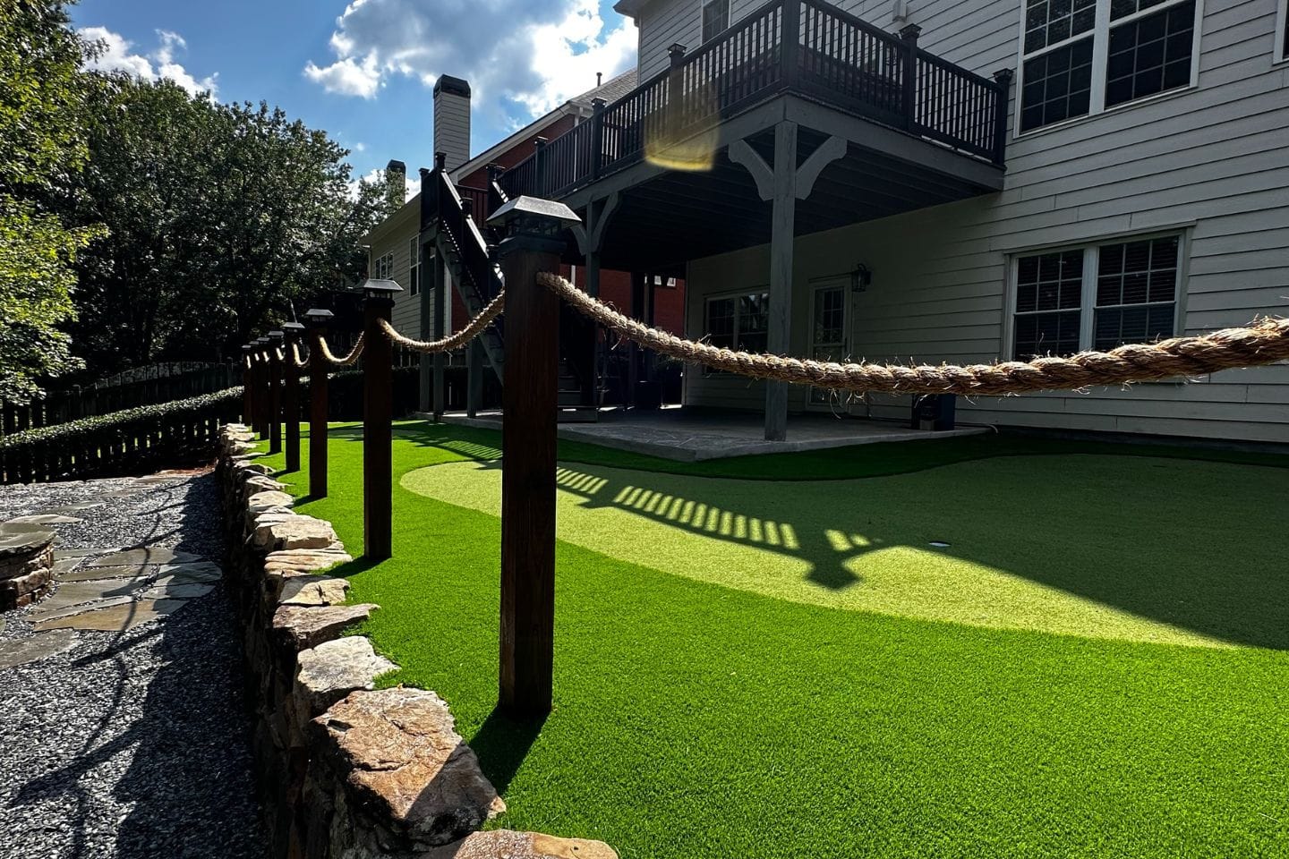 A backyard putting green with artificial grass, bordered by a stone pathway crafted by Atlanta hardscape contractors and wooden posts connected by rope. A deck overhead casts shadows on the green, while trees and clouds add depth to the background.