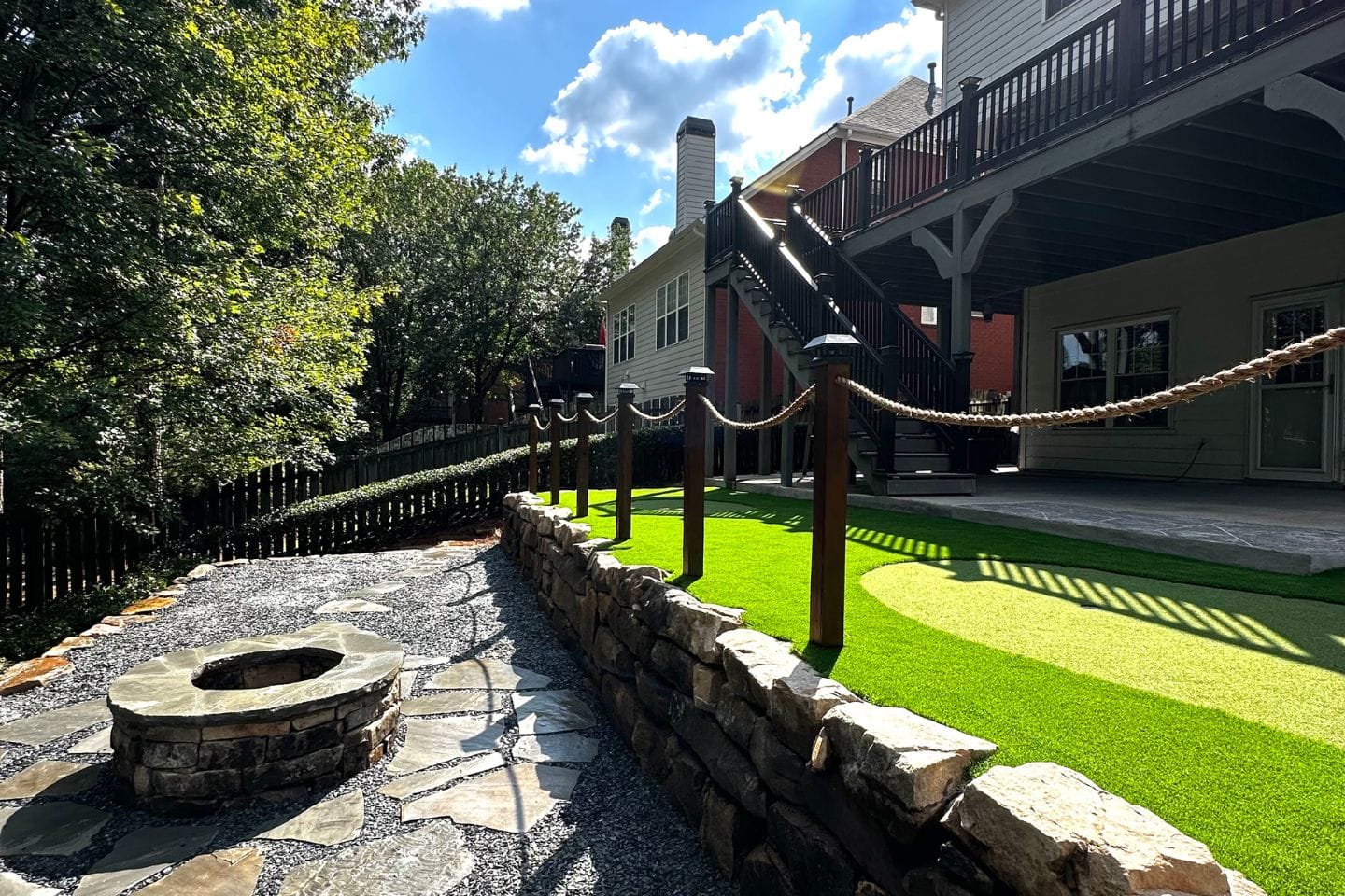 Our backyard boasts a sophisticated design with artificial grass, a stone patio, and a cozy fire pit crafted by top Atlanta hardscape contractors. A wooden rope fence adds charm, while a multilevel deck enhances the home. Lush trees stand against the backdrop of a cloudy sky.