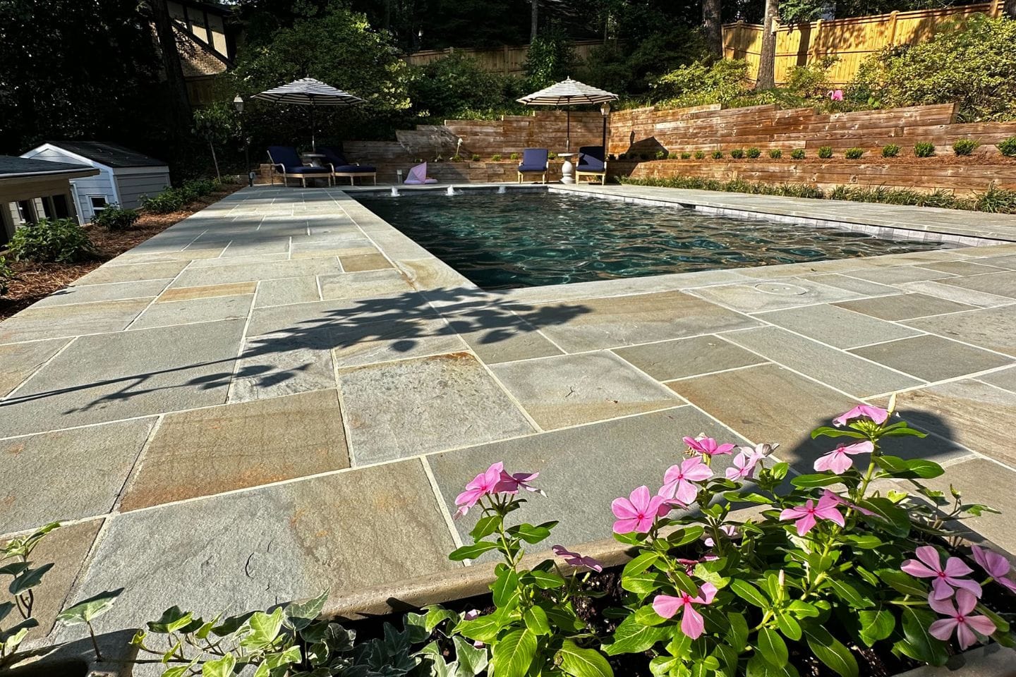 Stone patio with a rectangular pool, expertly crafted by Atlanta hardscape contractors, is surrounded by a lush garden. Several lounge chairs are shaded by umbrellas in the background, while vibrant pink flowers bloom in the foreground.