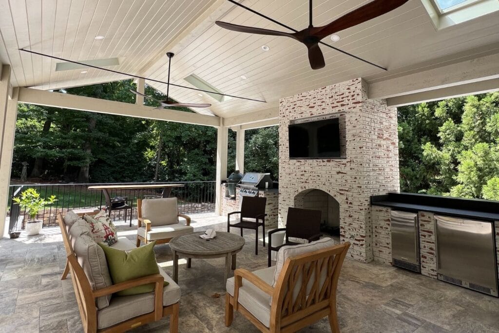 Atlanta hardscape contractors have crafted an outdoor patio with a cozy seating area, ceiling fans, a brick fireplace, TV, and built-in grill nestled near lush green trees.