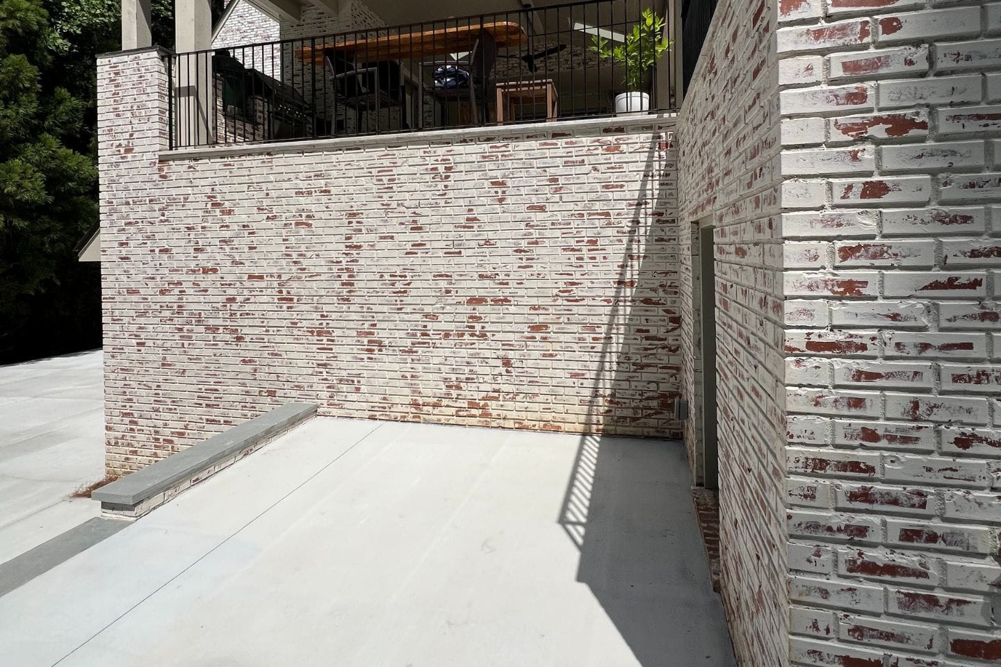 Amidst the sunlit, whitewashed brick exterior, crafted by Atlanta hardscape contractors, a patio above boasts a black railing and furniture in the background.