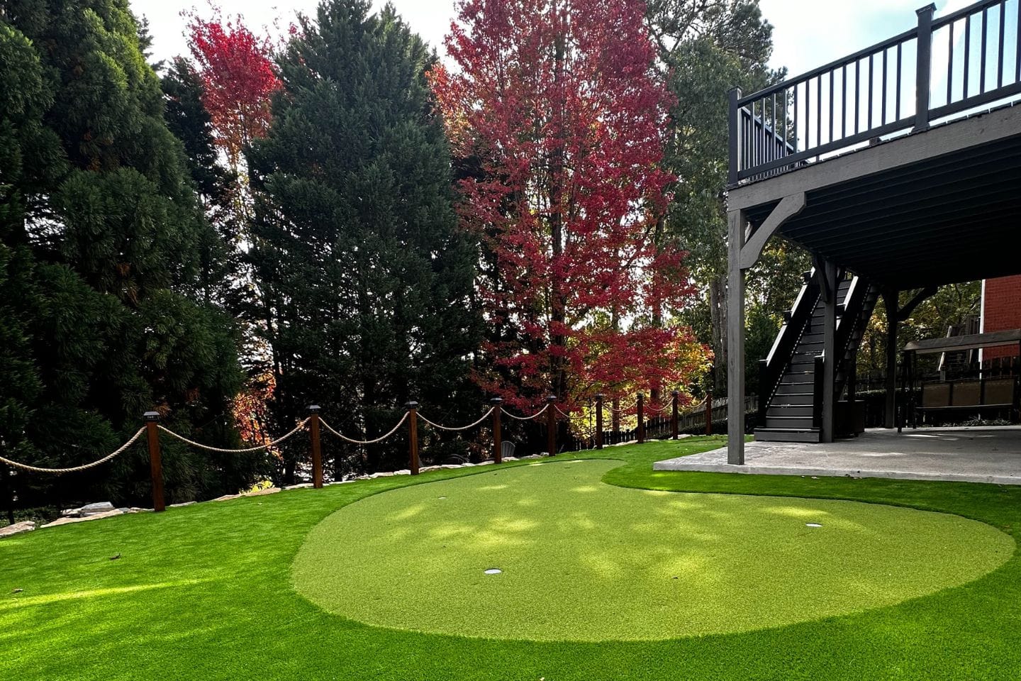 A backyard designed by Atlanta hardscape contractors features a lush green putting area surrounded by vibrant trees with red and green foliage, adjacent to a raised deck with stairs.