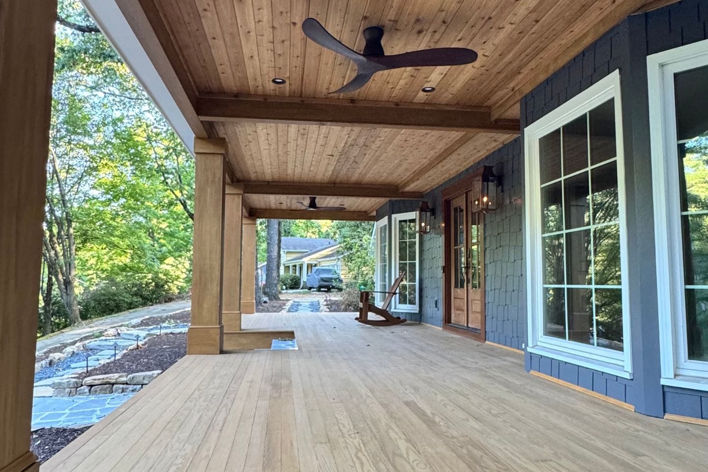 A spacious wooden porch with a ceiling fan and rocking chair invites relaxation. Large windows and a wood-paneled ceiling create an inviting space, crafted with care by Atlanta hardscape contractors. Trees frame the scene, adding to the charming backdrop of a nearby house.