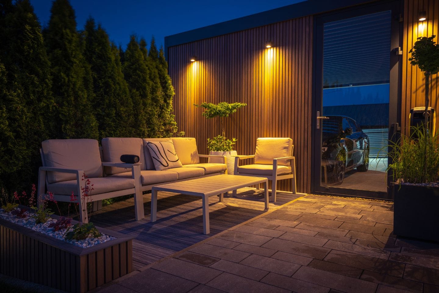 The outdoor patio at night features beige furniture, a table, and potted plants. Low-voltage lights elegantly illuminate the area, casting a warm glow. Through the glass door, a dark car is visible in the gentle ambiance.