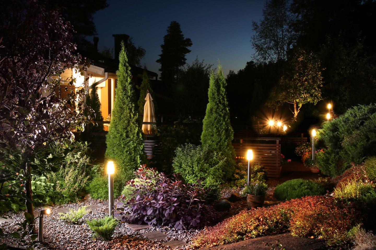 Illuminated garden pathway at dusk with trees, shrubs, and decorative plants. Low-voltage lights create a serene atmosphere near a house.