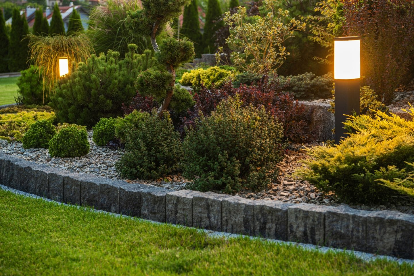 A landscaped garden with small shrubs and gravel features illuminated by two low-voltage lights at dusk.