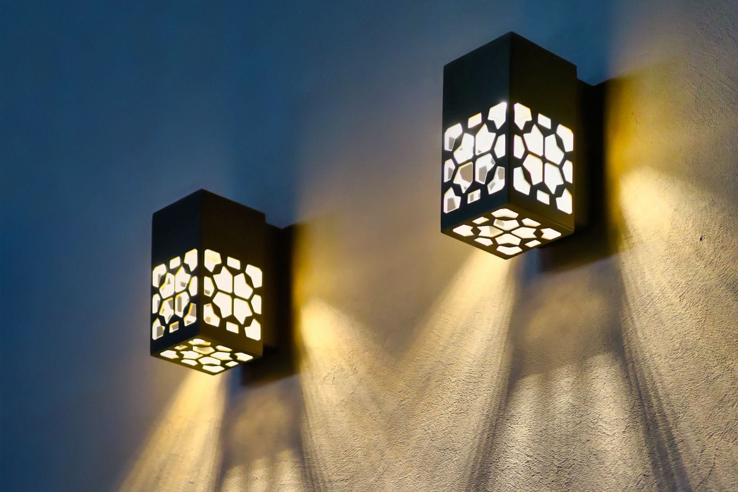 Two wall-mounted lanterns with geometric cutout patterns cast shadows on a textured wall, illuminated with warm, low-voltage lights.
