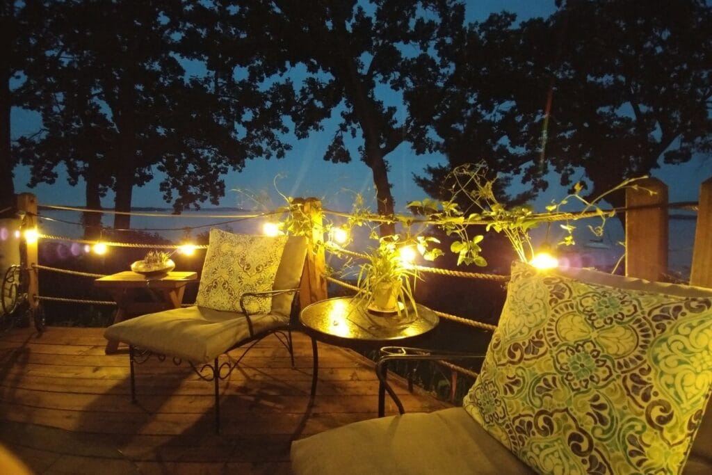A cozy outdoor seating area at dusk features warm landscape lighting, two cushioned chairs, a small table, plants, and a backdrop of trees.