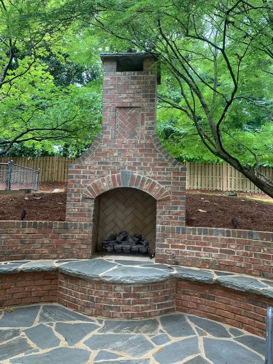 Home to an outdoor brick fireplace with a herringbone pattern, nestled against a fenced garden with lush green trees and flagstone flooring.