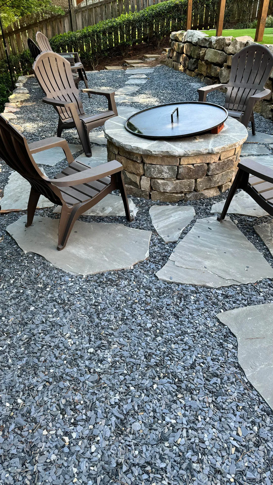 Patio with a stone fire pit surrounded by wooden Adirondack chairs on a gravel surface.