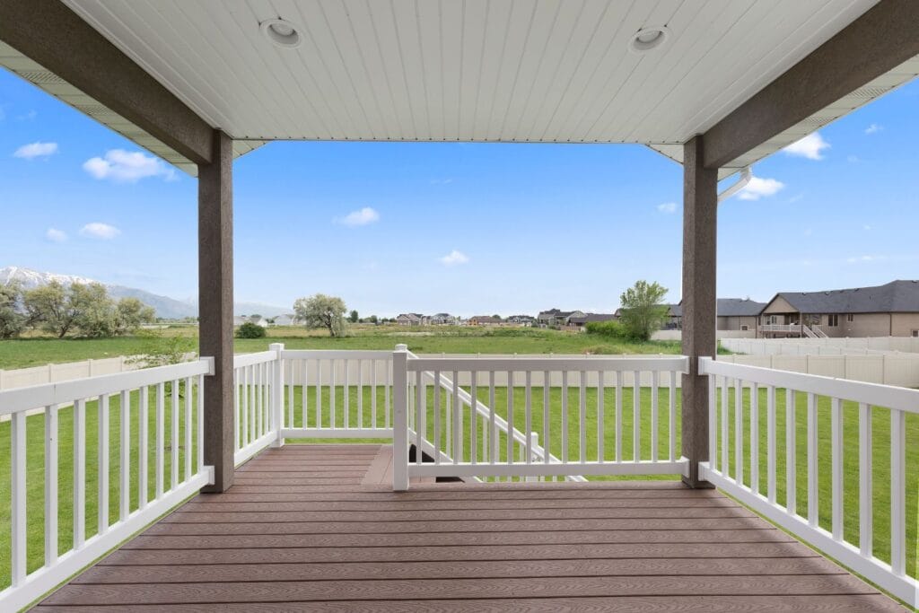 View from a covered deck featuring custom patio designs, overlooking an expansive green lawn, white fence, and distant houses under a clear blue sky.