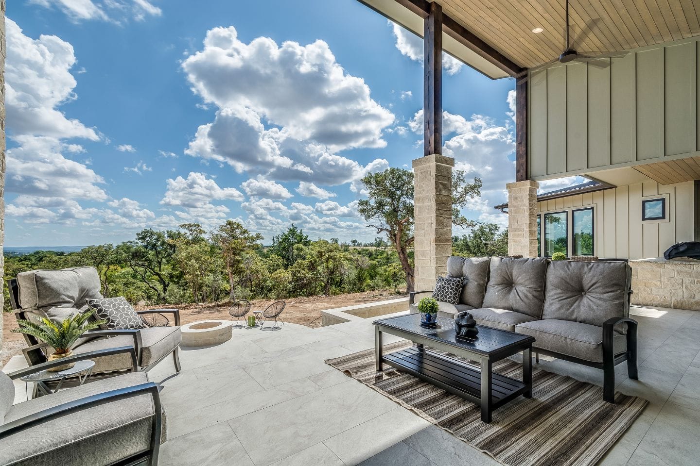 Discover custom patio designs featuring cushioned seating, a coffee table, and a rug. This outdoor space offers breathtaking views of trees and a cloudy sky, with a fire pit and additional seating in the background for ultimate relaxation.