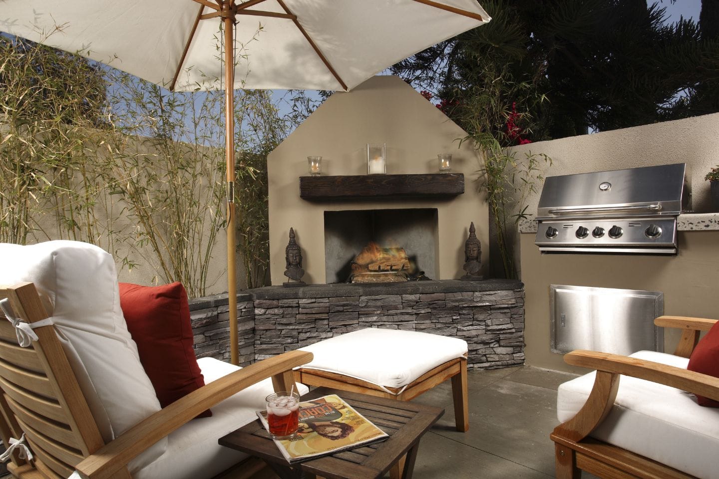 This custom patio design features cushioned wooden chairs, a small table with a drink and magazine, a stone fireplace, bamboo plants, a large umbrella, and a stainless steel grill.