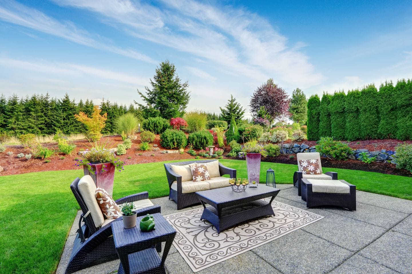 The patio features a wicker furniture set on a decorative rug, nestled in a manicured garden with trees and shrubs beneath the clear blue sky. The space showcases custom patio designs, adding unique charm to this serene oasis.