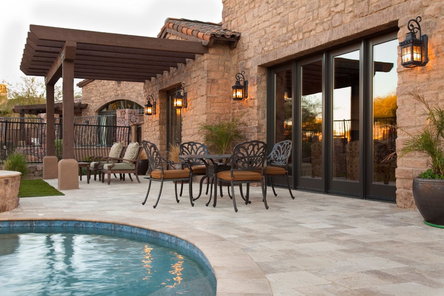 A custom patio design features a pergola, circular table, four chairs, and outdoor lighting beside a swimming pool, set against the stone facade of a house with large windows.