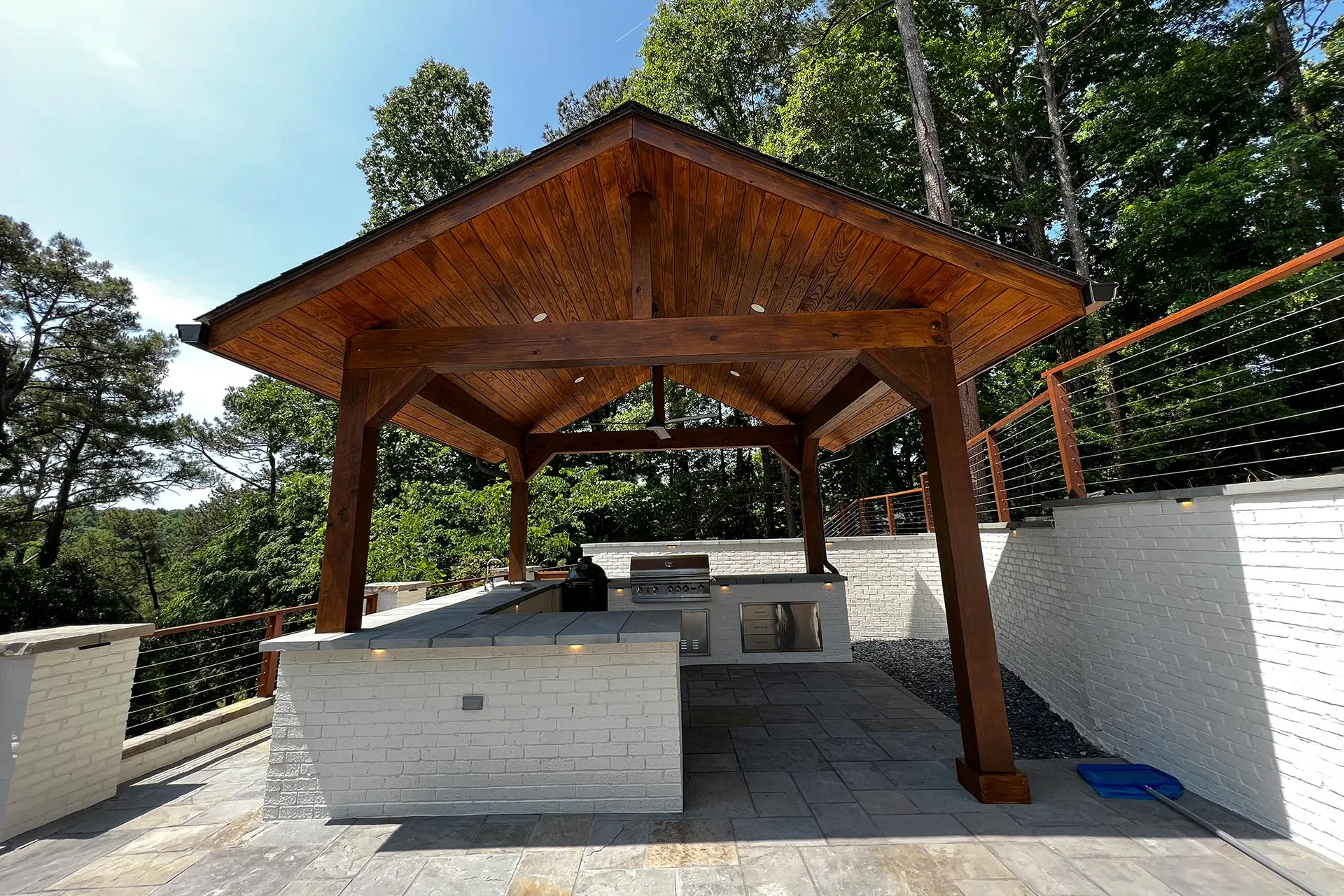 A wooden pavilion with a pitched roof shelters an outdoor kitchen featuring a grill and countertops, crafted by renowned Atlanta hardscape contractors. White brick walls and lush trees provide the perfect backdrop for this serene oasis.