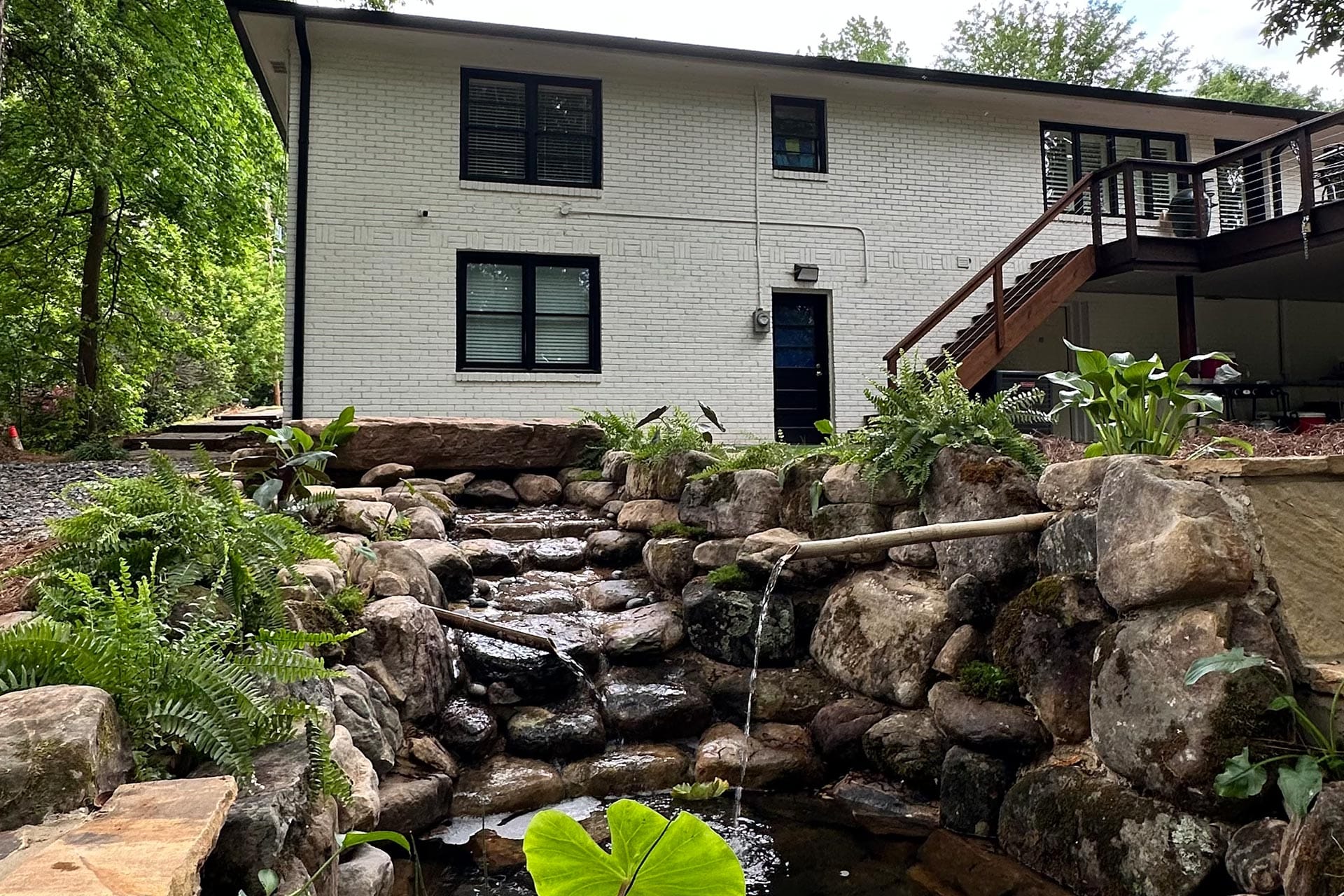A two-story white brick house with a wooden deck overlooks a stone waterfall feature, masterfully crafted by Atlanta hardscape contractors, and is surrounded by lush greenery.