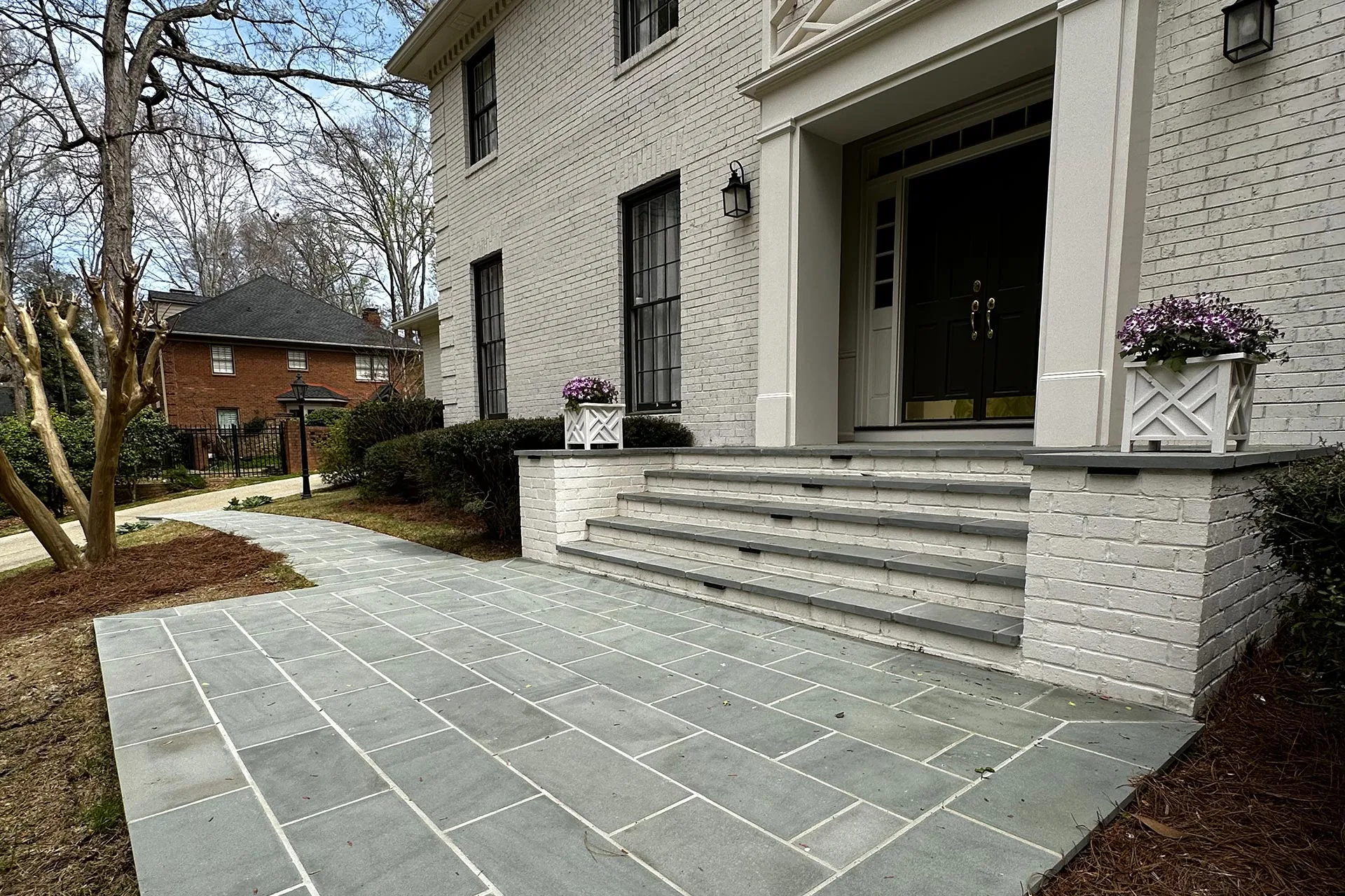 A beautifully crafted stone walkway, reminiscent of Atlanta hardscape contractors' finest work, leads to the front steps of a brick house with green double doors. Flanked by white planters filled with vibrant purple flowers, the entrance is elegantly framed by lush trees and shrubs.