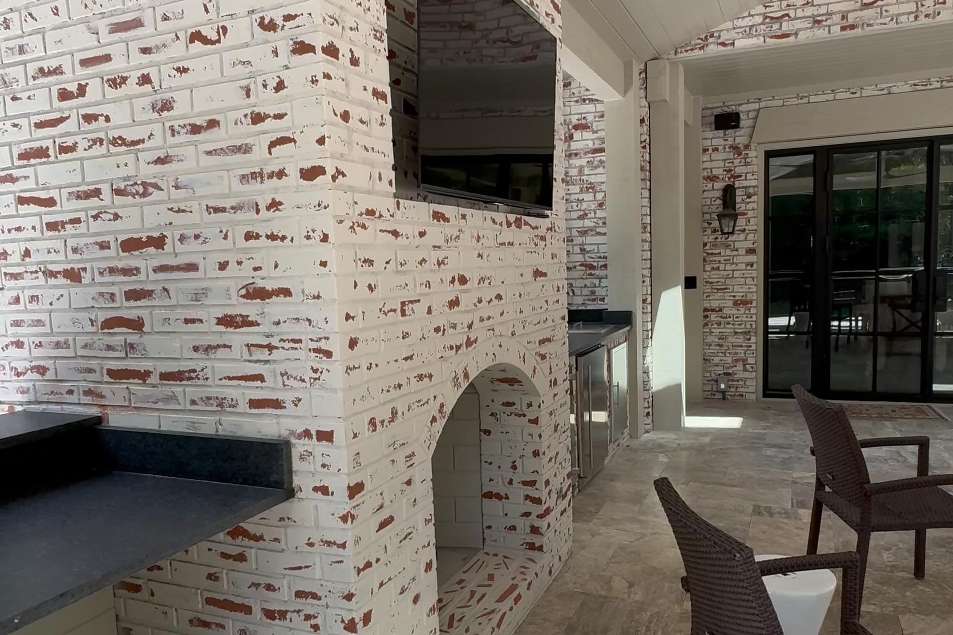 The outdoor kitchen area features whitewashed brick walls and an archway, impeccably crafted by Atlanta hardscape contractors. Wicker chairs complement the sleek countertop, with a closed black door and a mounted TV screen adding modern touches.