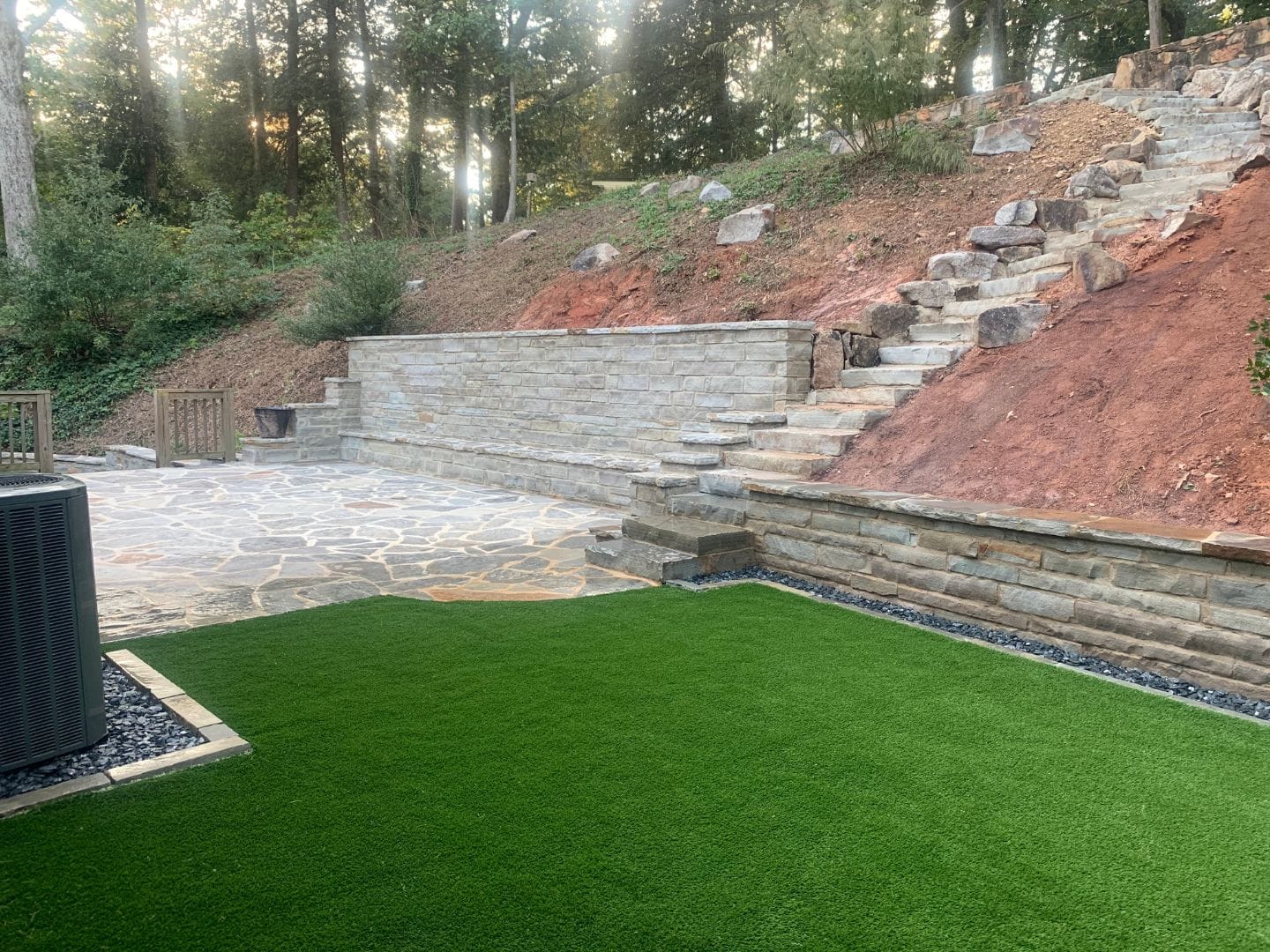 A landscaped backyard with artificial grass and beautifully designed patios, featuring a stone path and a rock staircase leading up to an elevated garden area surrounded by trees and plants.