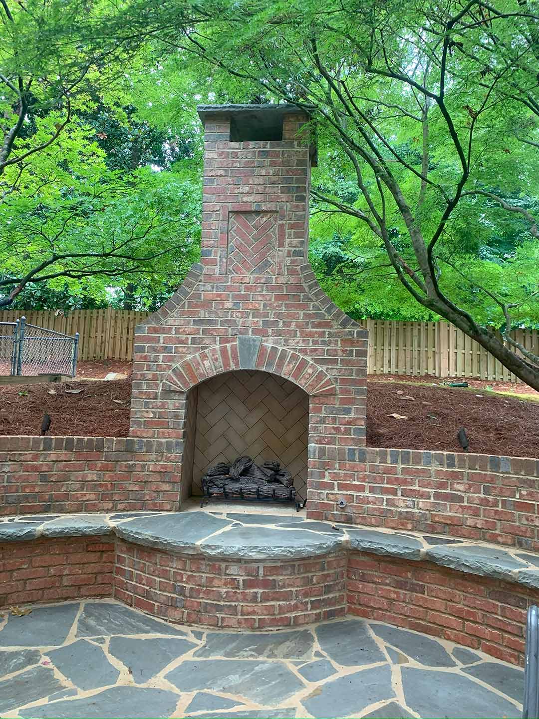 Outdoor brick fireplace with a herringbone patterned interior, surrounded by a stone patio and a wooden fence in a garden setting. Trees and greenery are visible in the background, making it an ideal spot for backyard heating.