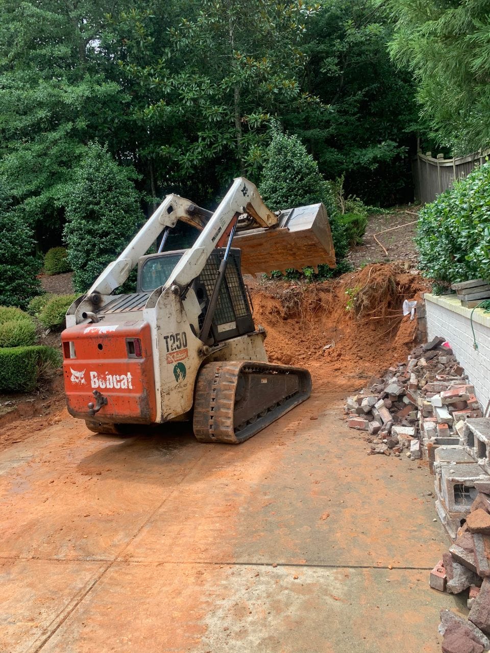 A Bobcat T250 compact track loader is performing excavation work, moving dirt and debris in a residential backyard. Piles of soil and dislodged bricks are visible around the work area, preparing the site for new retaining walls.