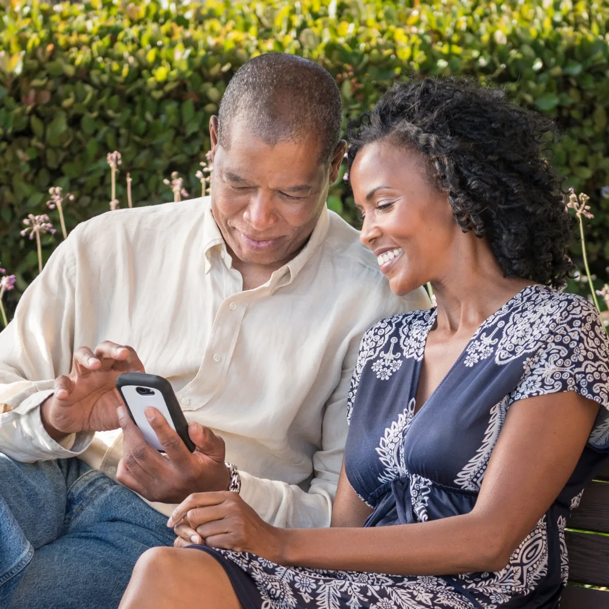 A man and woman sit on a bench outdoors, surrounded by lush greenery. The are looking at Atlanta Hardscape Kings website, getting ready to call them about doing some hardscaping for their yard.