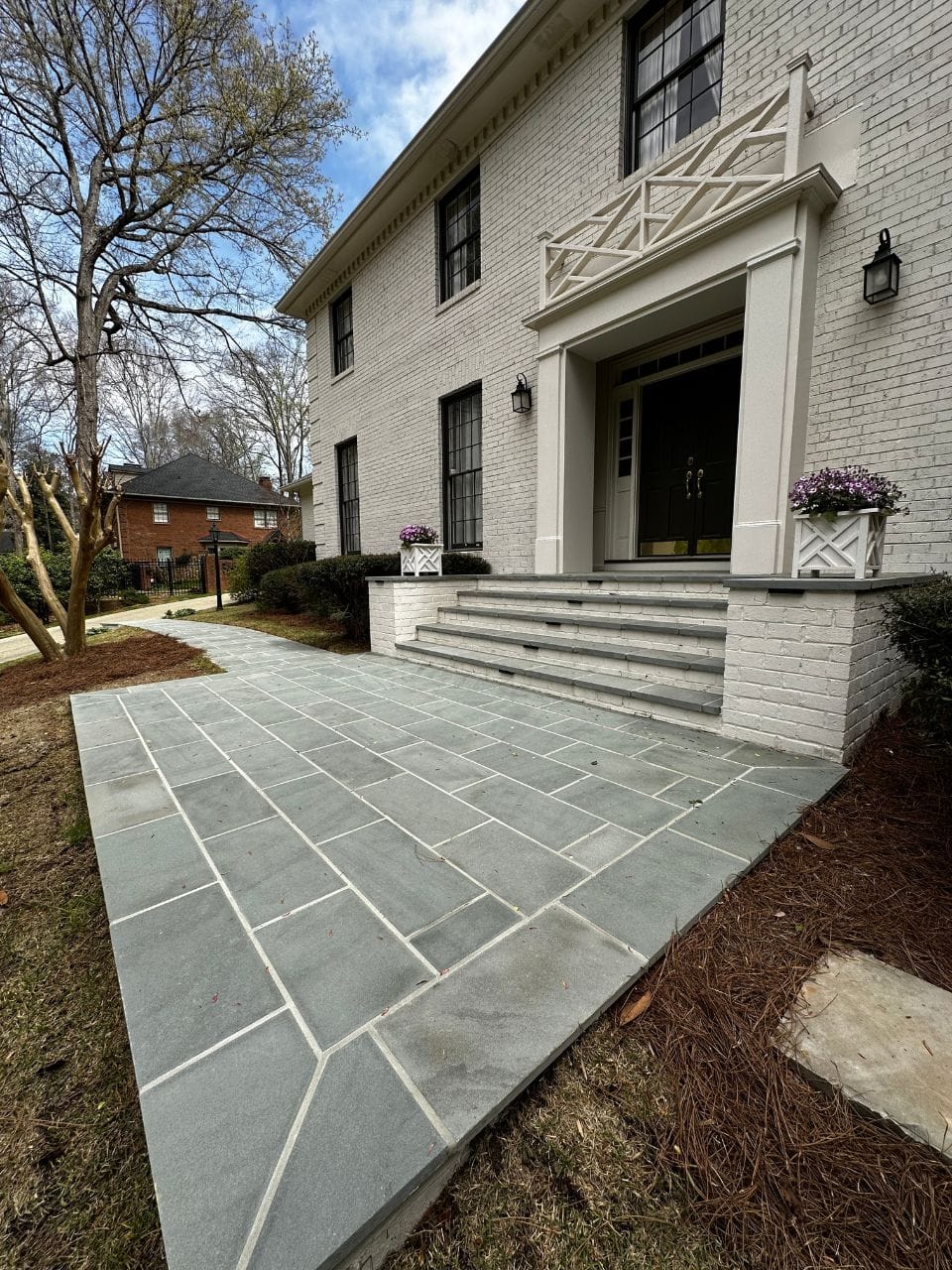 A front view of a brick house with a gray stone-tiled pathway leading to a raised entrance with steps, flanked by potted flowers, and adjoining cozy patios perfect for relaxing outdoors.