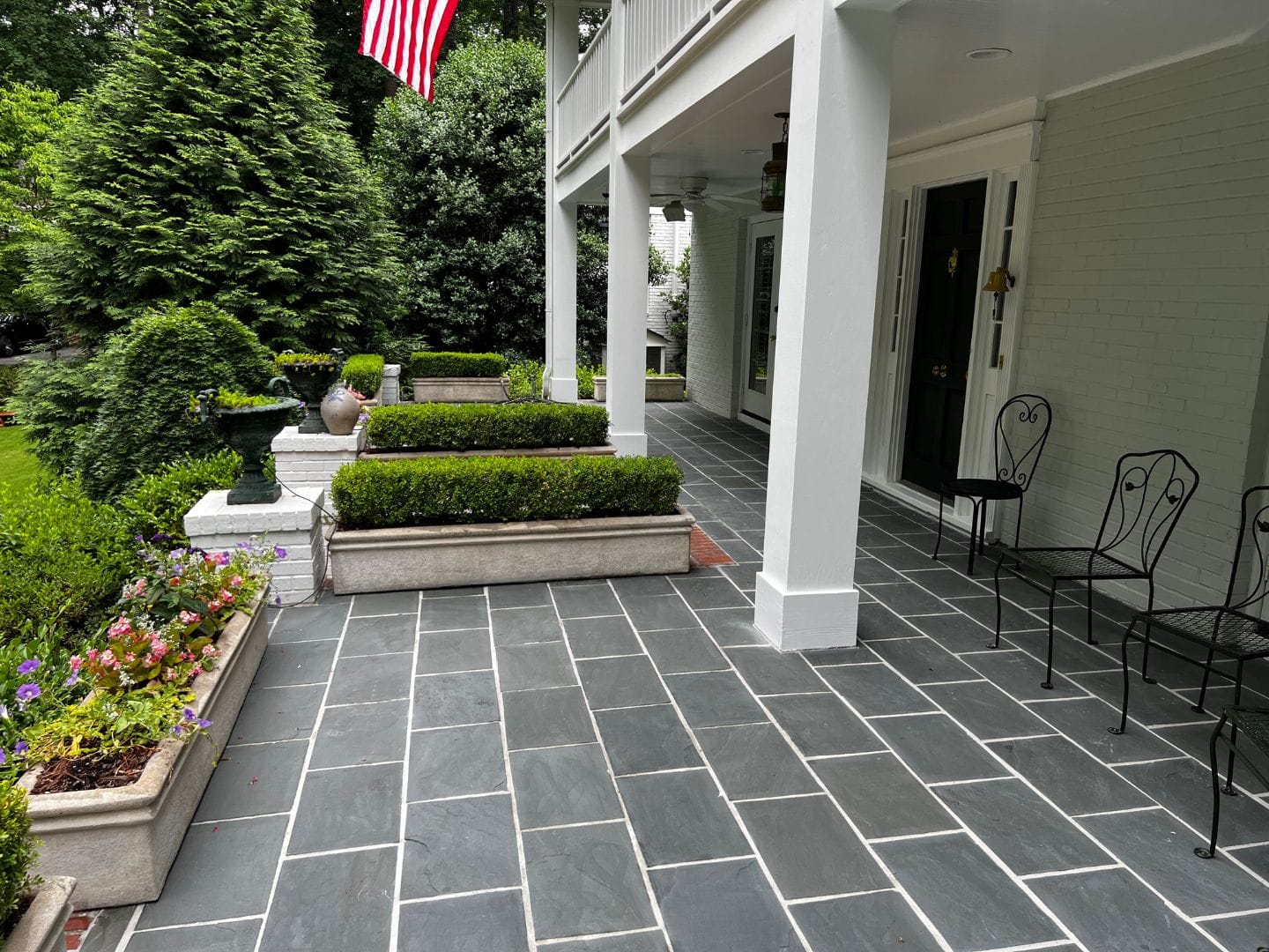 A porch with gray slate flooring, an American flag, black metal chairs, potted plants, and lush green bushes brings a serene charm reminiscent of cozy patios.