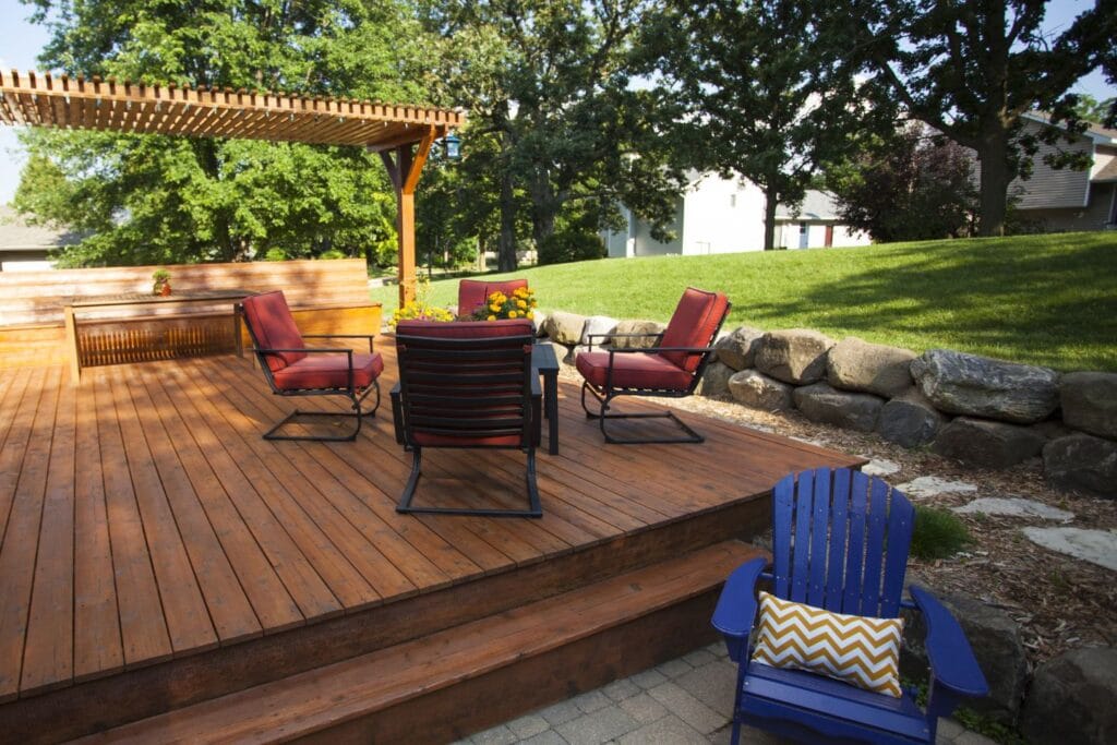 The wooden deck boasts four red-cushioned chairs circling a cozy fire pit, complemented by a blue chair with a pillow. To the left, a pergola adorned with plants enhances this charming outdoor space, while a rock wall elegantly borders the lush grass.