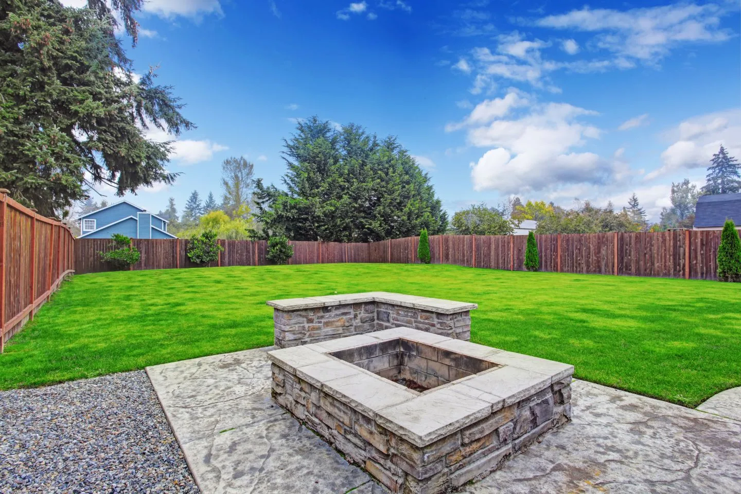 A spacious backyard with a stone fire pit in the center, surrounded by a green lawn and a wooden fence, features an inviting deck under a clear blue sky with scattered clouds.
