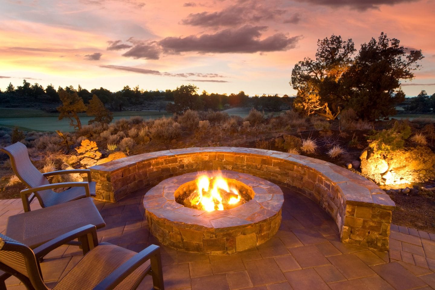 Outdoor patio with a curved stone fire pit and two chairs, seamlessly connecting to an adjacent deck. The sky displays a colorful sunset with silhouetted trees and shrubs in the background.