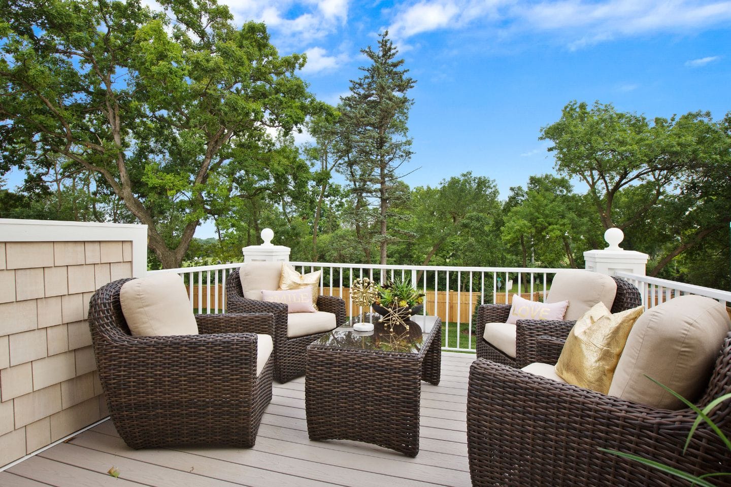 The outdoor seating area features wicker chairs and a table on a spacious deck, nestled among trees under a clear blue sky.