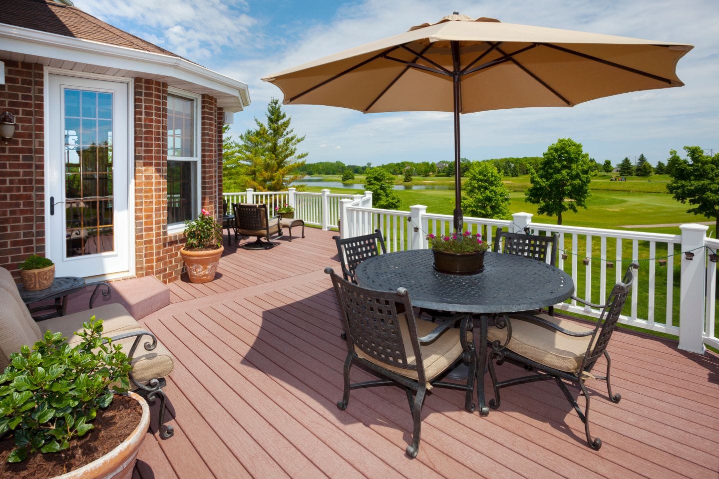 A spacious outdoor deck features a round table, chairs, and an umbrella. Potted plants complement the view of green landscapes with trees under a blue, partly cloudy sky. This inviting deck is perfect for enjoying nature's tranquility.