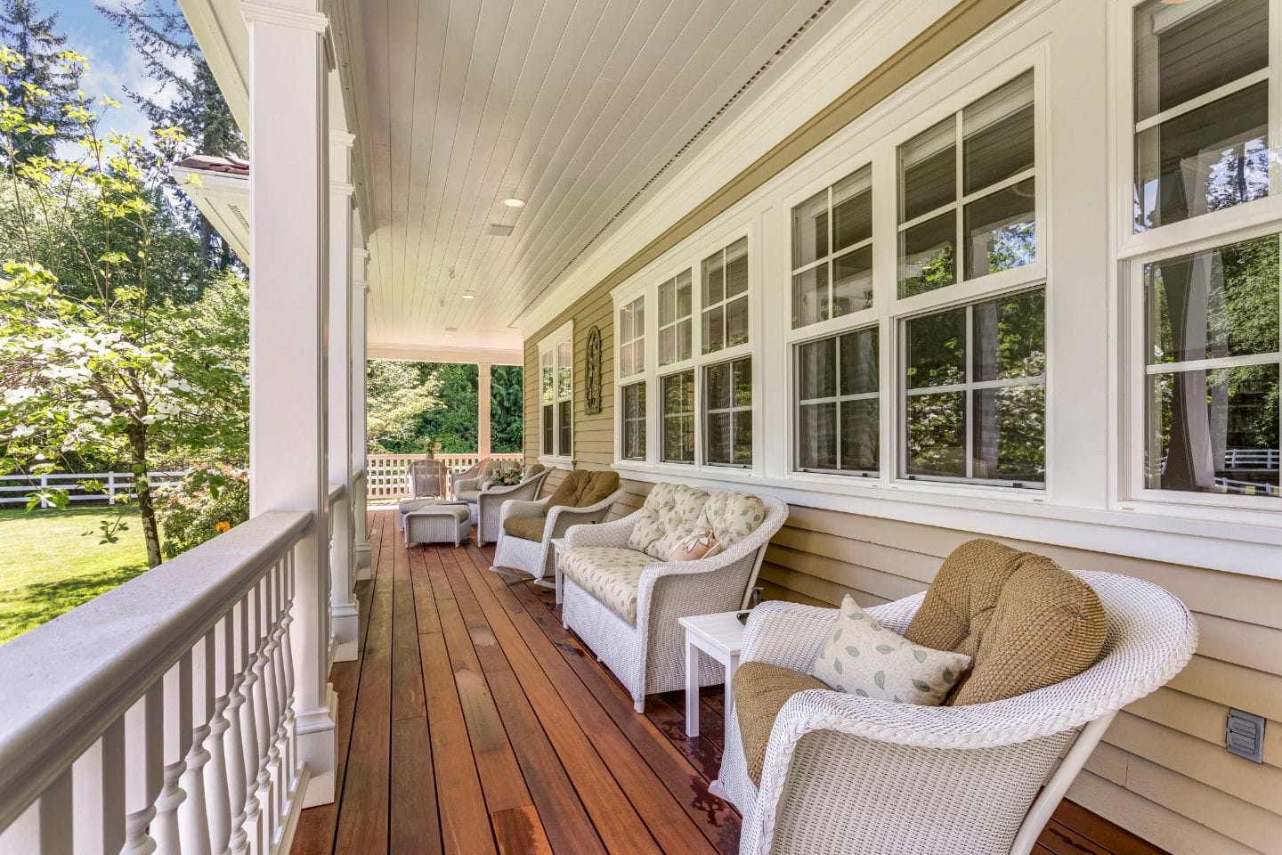 A covered porch with wooden flooring seamlessly connects to adjacent decks, featuring white wicker chairs and a sofa with cushions. Large windows line the wall, offering a view of the green, tree-filled yard beyond.
