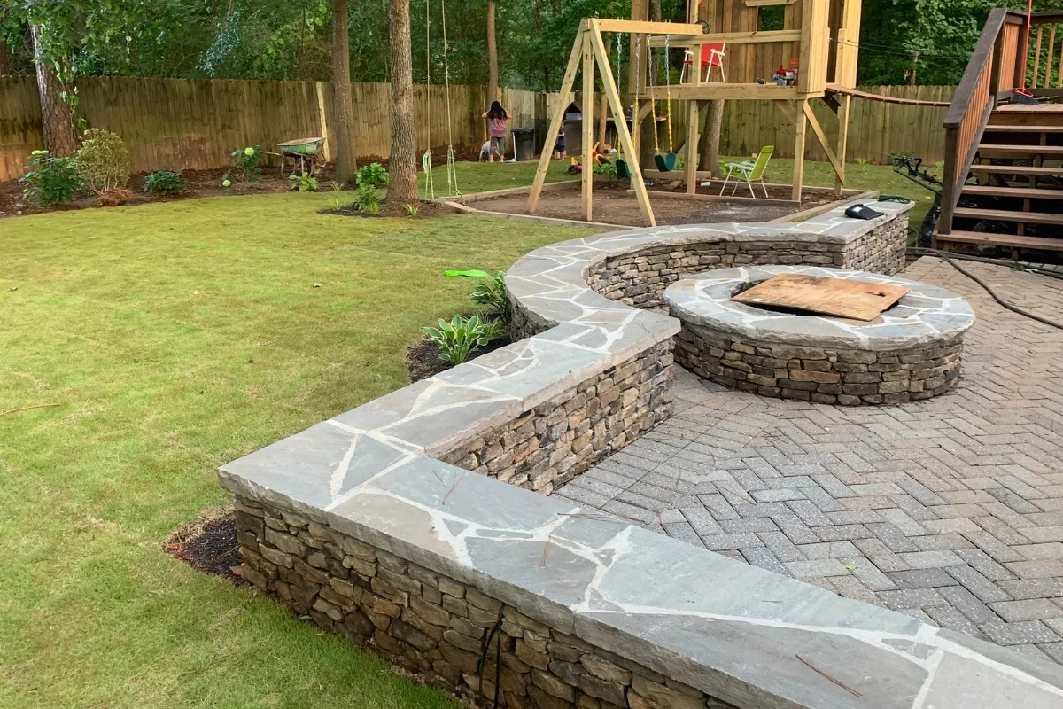 A backyard features a stone retaining wall and seating area with a fire pit, a swing set, a treehouse, and a patch of green grass with a wooden fence in the background. Tools and gardening equipment are visible.