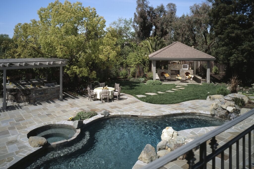 Backyard with a swimming pool, stone tiles, a shaded outdoor seating area, a dining area with table and chairs, and lush greenery—including several trees and a lawn—enhanced by cozy outdoor fireplaces for added warmth.